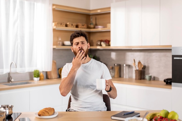 Sleepy tired bored adult caucasian man in white tshirt enjoys cup of coffee yawns and drinks cup of coffee