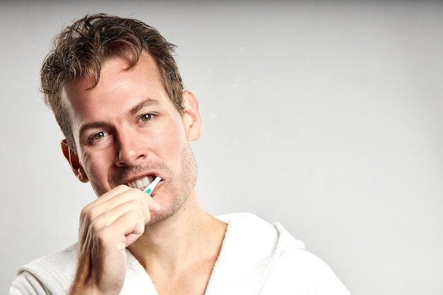 Sleepy tired caucasian american guy brushing tooth in the morning isolated on gray wall background studio portrait Skin care healthcare cosmetic procedures concept Mockup copy space