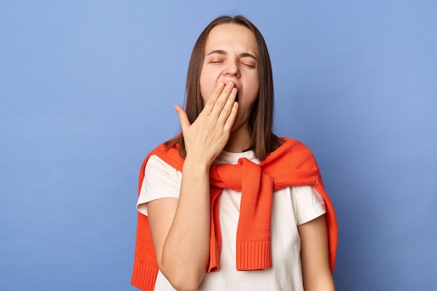 Sleepy woman wearing white Tshirt and sweater tied over shoulders yawns as being tired and needs good rest after hard working day gets up early in morning standing isolated on blue background