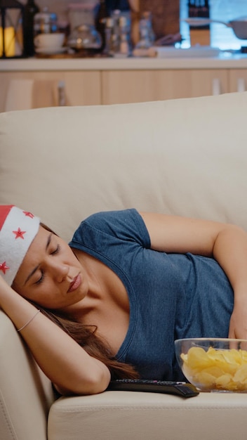 Sleepy woman with santa hat watching television on sofa. Festive adult falling asleep while looking at TV screen with TV remote control and bowl of chips. Person sleeping on christmas eve.