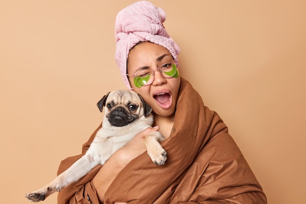 Sleepy young European woman yawns winks eye keeps mouth opened wears towel wrapped in blanket holds pug dog winks eye isolated over brown studio background Female model with pedigree pet indoors