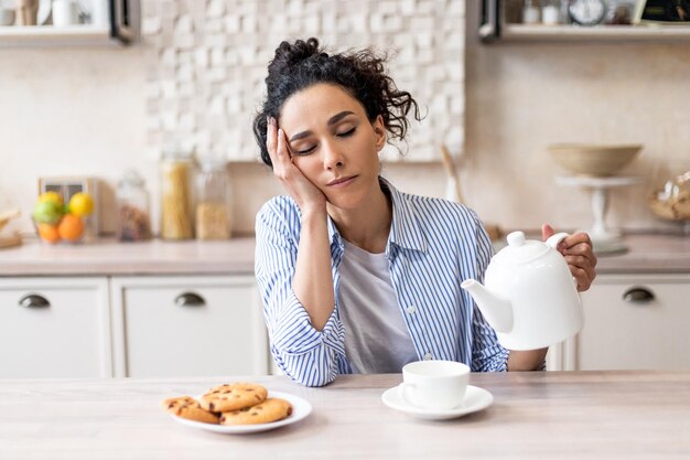 眠そうな若い女性がカップにお茶を注いで、キッチンのテーブルに座って早起きして寝ている