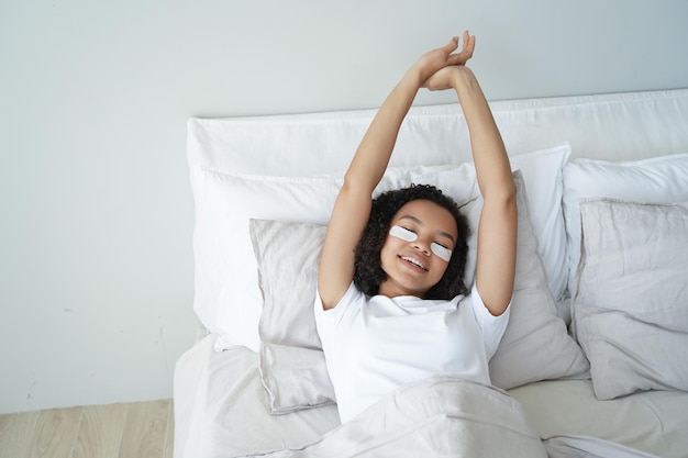 Sleepy young mixed race girl with under eye patches stretching\
lying in bed skincare wellness