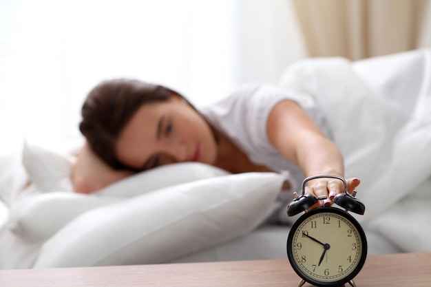 Sleepy young woman stretching hand to ringing alarm willing turn it off. Early wake up, not getting enough sleep, getting work concept.