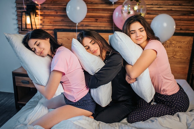 Photo sleepy young women sitting on bed in room. their head lying on pillows they have in hands. they sleep. women smile. they sit one after another.