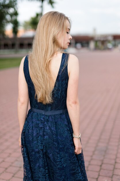 Slender blonde girl in dark-blue dress turned her back looking away on pavement.