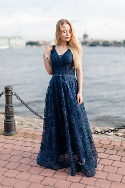 Slender blonde girl in elegant dark-blue floor-length dress on embankment mysteriously looked down.