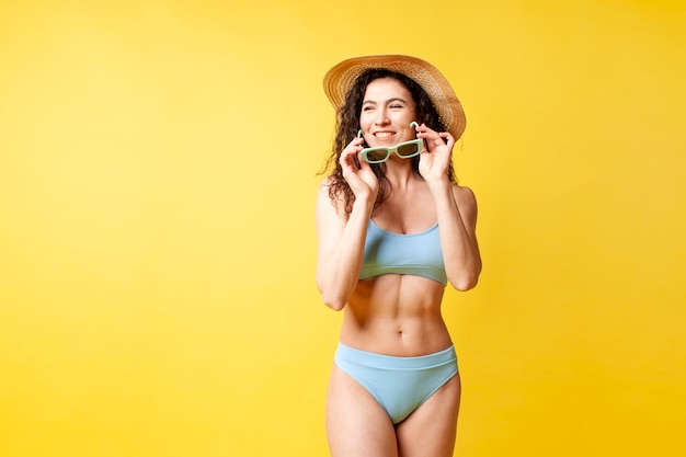Photo slender brunette girl in blue swimsuit and with curly hair holds sunglasses on yellow background