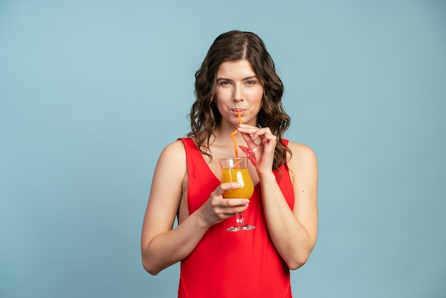 Slender girl on a blue background drinks orange juice on a background of blue wall