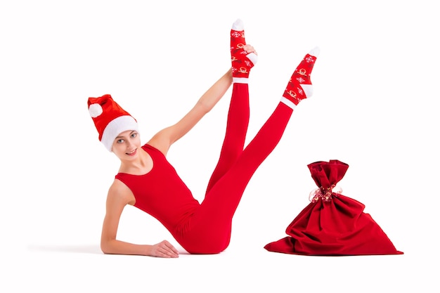 Slender girl gymnast in red tight-fitting suit and santa claus hat posing next to a bag for gifts isolated on white background