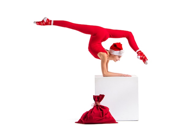Slender girl gymnast in red tight-fitting suit and santa claus hat posing next to a bag for gifts isolated on white background