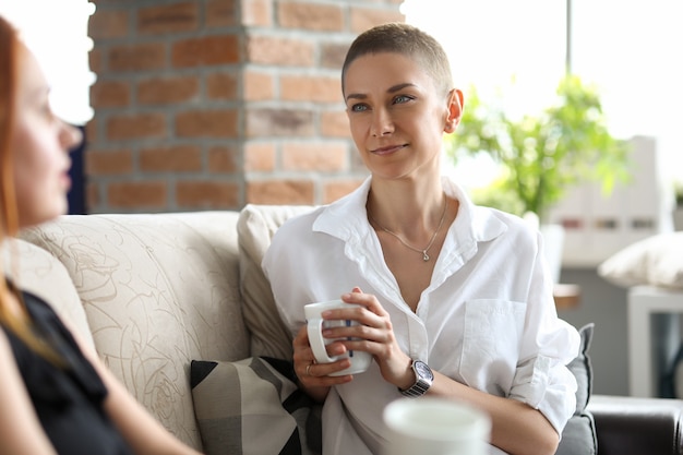 A slender girl in a white blouse sits on a sofa and drinks coffee with her friend. Creative young woman resting in office during lunch