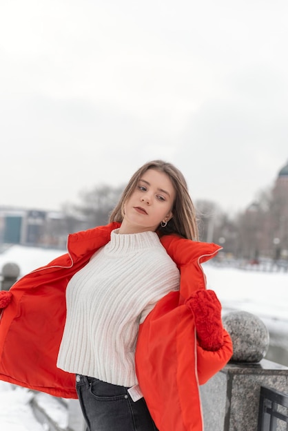 Slender girl in white knitted sweater and red short down jacket with knitted mittens poses in snowcovered park