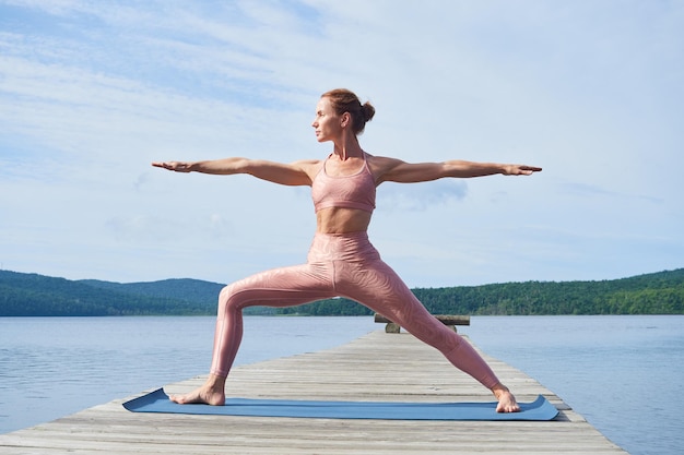 Una donna matura snella e forte in una tuta rosa pratica lo yoga in riva al mare durante l'estate