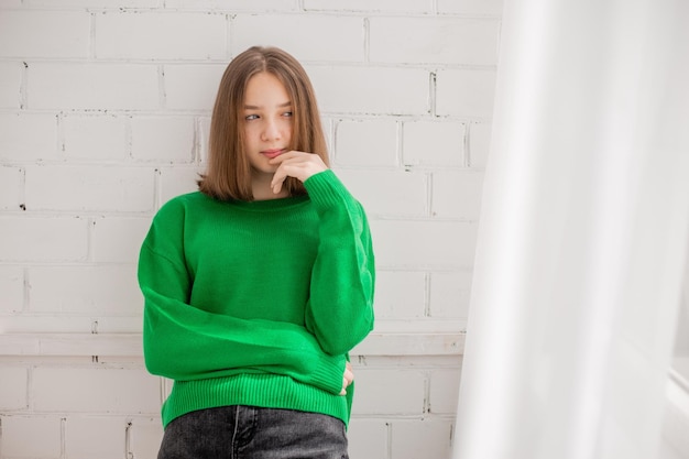 Стройная девочка-подросток в джинсах и зеленом свитере позирует на фоне белой кирпичной стены. естественная красота, без макияжа, естественное освещение. модная съемка. место для текста. Фото высокого качества