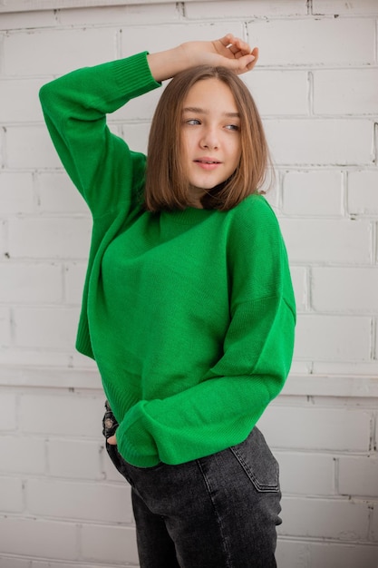 Slender teenage girl in jeans and a green sweater poses against a white brick wall. natural beauty, no makeup, natural lighting. fashion shooting. space for text. High quality photo