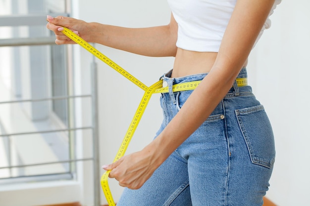 Slender woman in jeans and a white T-shirt measures her waist.