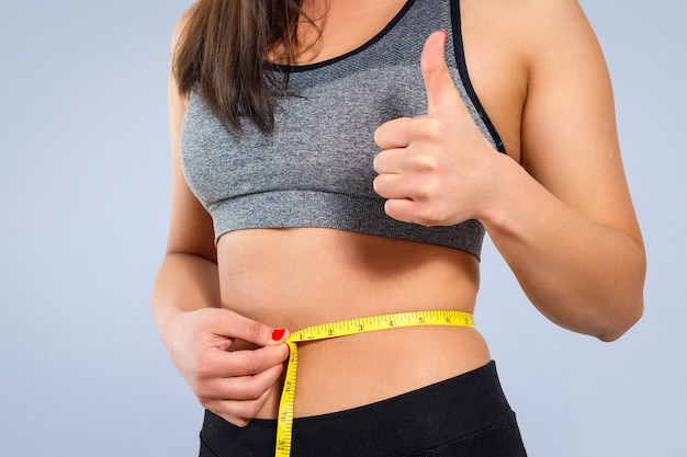 A slender woman measures her waist with a measuring tape. Against a yellow background close-up.