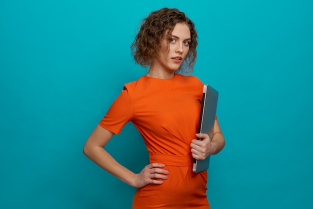 Slender woman in orange dress holding laptop.