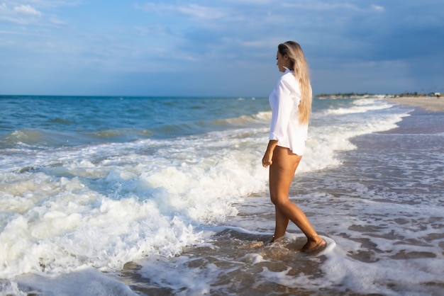 A slender young girl in a gentle blue swimsuit and a white shirt, walks along a wide sandy beach near the blue sea with white waves