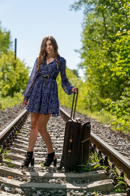 Foto una giovane ragazza snella con un vestito leggero è in piedi sui binari aggrappati al lungo manico di una valigia da viaggio