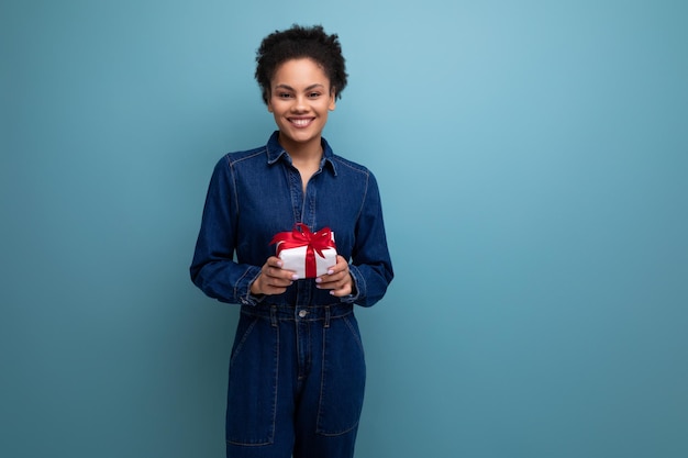 A slender young hispanic brunette woman with fluffy curly hair in a blue denim suit hugging a gift