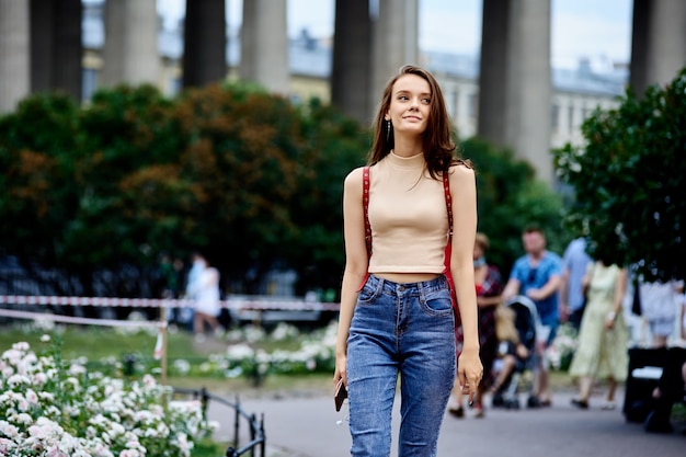 Slender young woman in jeans and tank top walks around city on summer evening