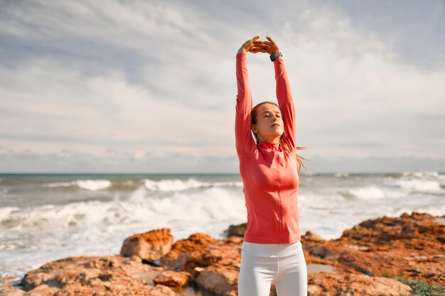 A slender young woman in sportswear doing exercises while standing on the stones against the background of the sea. sports and recreation