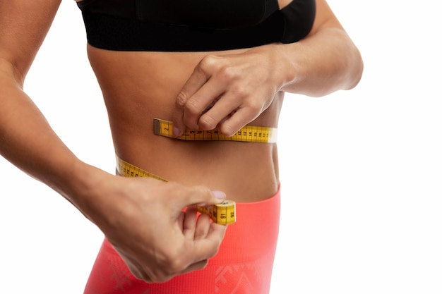 A slender young woman in sportswear measures her waist with a centimeter tape. Close-up. Isolated on white wall. Space for text.