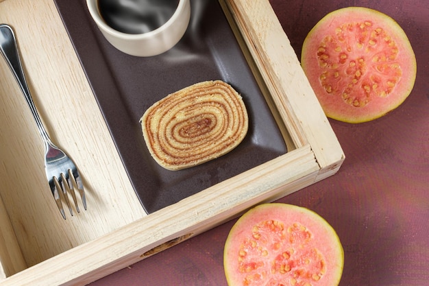 Slice of bolo de rolo on a brown plate, surrounded by guava, cup of coffee and a fork.