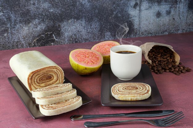 Slice of bolo de rolo surrounded by guava, cup and coffee beans on a red background (side view).