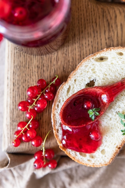 Photo slice of bread and red currant jam or jelly in a spoon, wooden cutting board. top view.