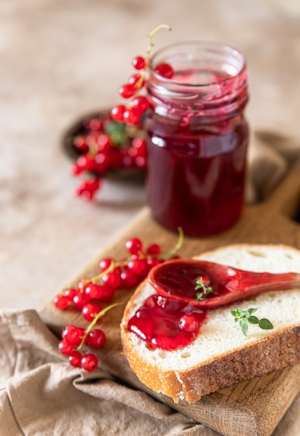 Slice of bread and red currant jam or jelly in a spoon, wooden cutting board.