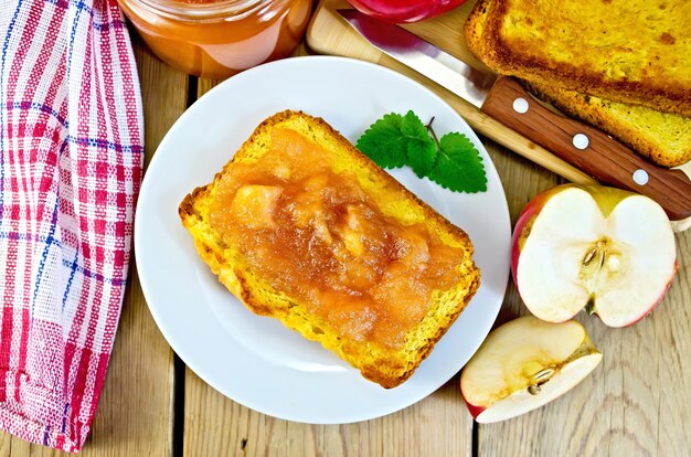 Slice of bread with apple jam and mint on a plate, slices of apple, a jar of jam, napkin, knife on background wooden plank