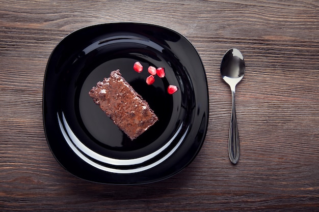 Slice of brownie on black plate on a wooden table with a spoon and pomegranate seeds