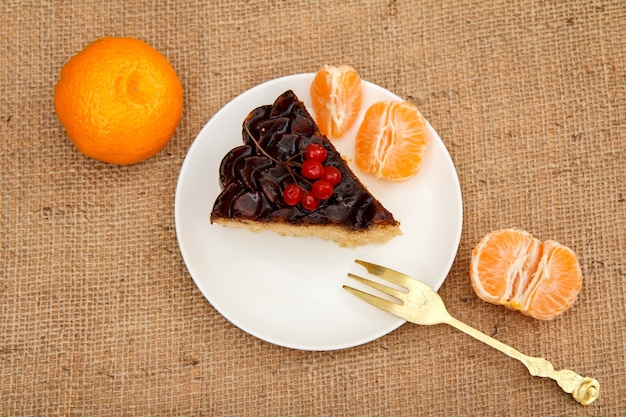 Slice of chocolate cake decorated with bunch of viburnum, fork on plate and oranges on table with sackcloth. Top view.