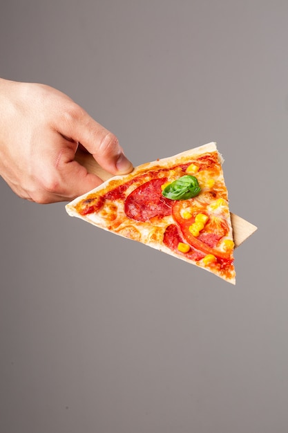 Slice of Delicious hot homemade pizza in hand on gray background. Pizza - Fresh homemade pizza with pepperoni, cheese and tomato sauce, tomatoes, sweet corn and basil with copy space.