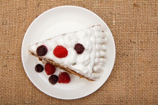 Slice of homemade biscuit cake decorated with whipped cream and raspberries on table with sackcloth. Top view