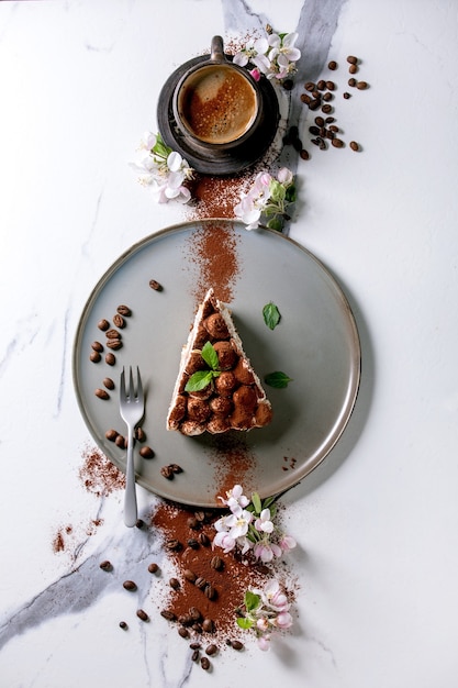 Slice of homemade gluten free tiramisu traditional italian dessert sprinkled with cocoa powder, blooming apple tree, coffee, mint leaves, coffee beans over white marble surface. Top view, flat lay