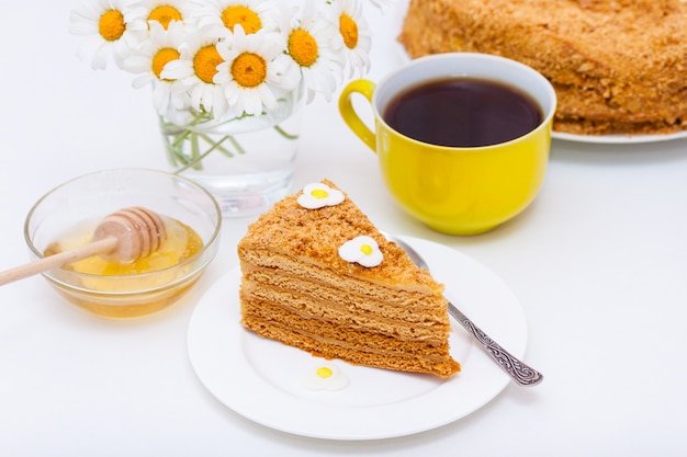 Slice of homemade layers honey cake with yellow cup of tea or coffee and chamomiles.