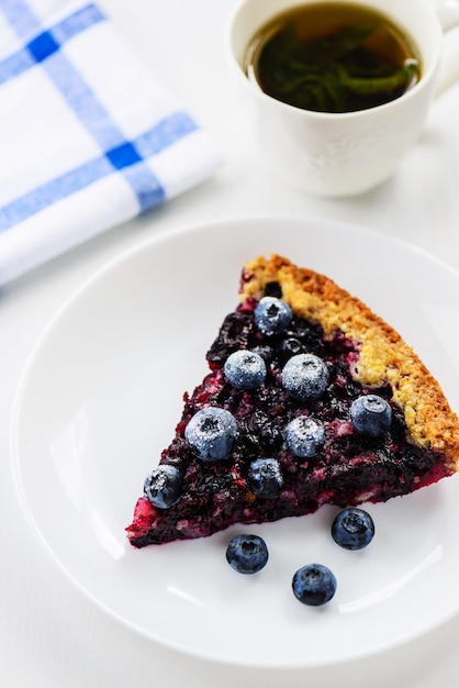 A slice of homemade pie with blueberries and a cup of green tea on a white table.