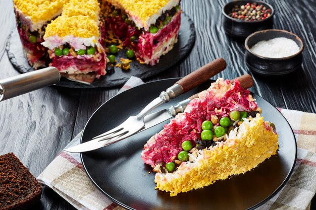 Una fetta di insalata di torta a strati con verdure, uova sode, prugne, noci tritate e carne di pollo sminuzzata servita su un piatto nero su un tavolo di legno nero, vista dall'alto, primo piano