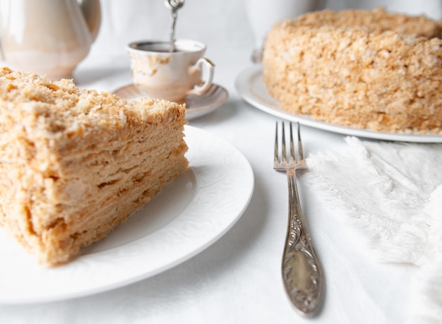 Fetta di torta napoleone multistrato con crema al burro su un piatto bianco primo piano accanto a un piatto c'è un tovagliolo e una forchetta sullo sfondo c'è una tazza teiera e un vaso di fiori sfondo bianco