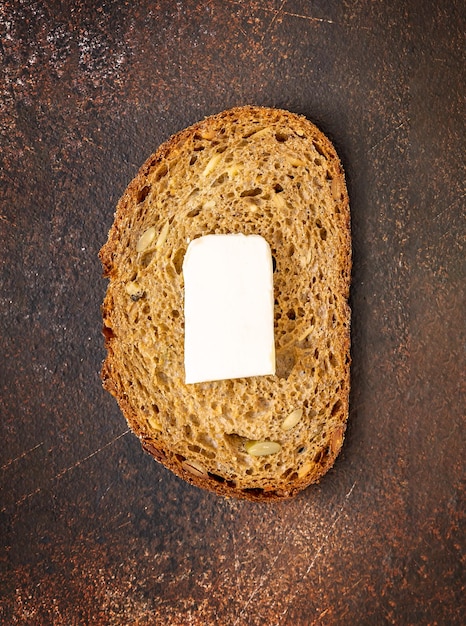 A slice of natural dark rye bread with a piece of fresh butter on a brown background