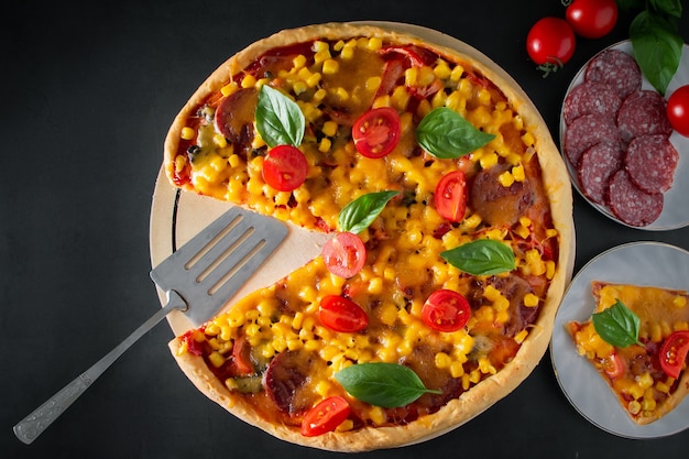 Slice of pizza with tomatoes and basil on a black background. National Italian food, pizza with tomatoes, corn, green basil and cheese on a round board. Vertical photo, place for text.