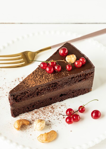 A slice of raw vegan chocolate cashew cake on a white plate,\
close-up view. white wooden background, red currants and nuts.