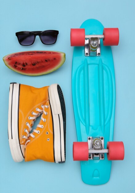 Slice of ripe watermelon and hipster youth accessories, cruiser board on blue background. Summer fun. Top view