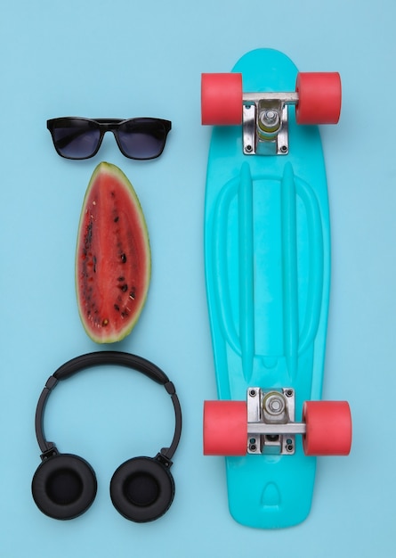 Slice of ripe watermelon and hipster youth accessories, cruiser board on blue background. Summer fun. Top view