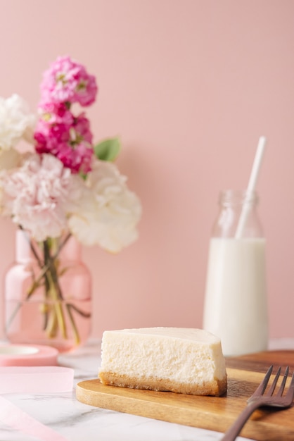 분홍색 배경에 꽃과 밀크가 있는 맛있는 홈메이드 치즈케이크 한 조각. 건강한 유기농 여름 디저트 파이 측면 보기 수직