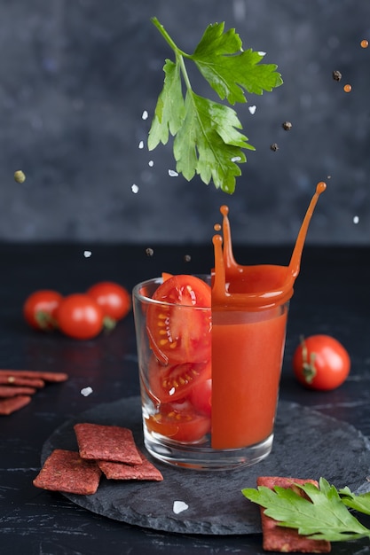 A slice of tomato drops in a glass of tomato juice, spray juice, fresh sliced tomato and bread rolls with tomato on gray background.